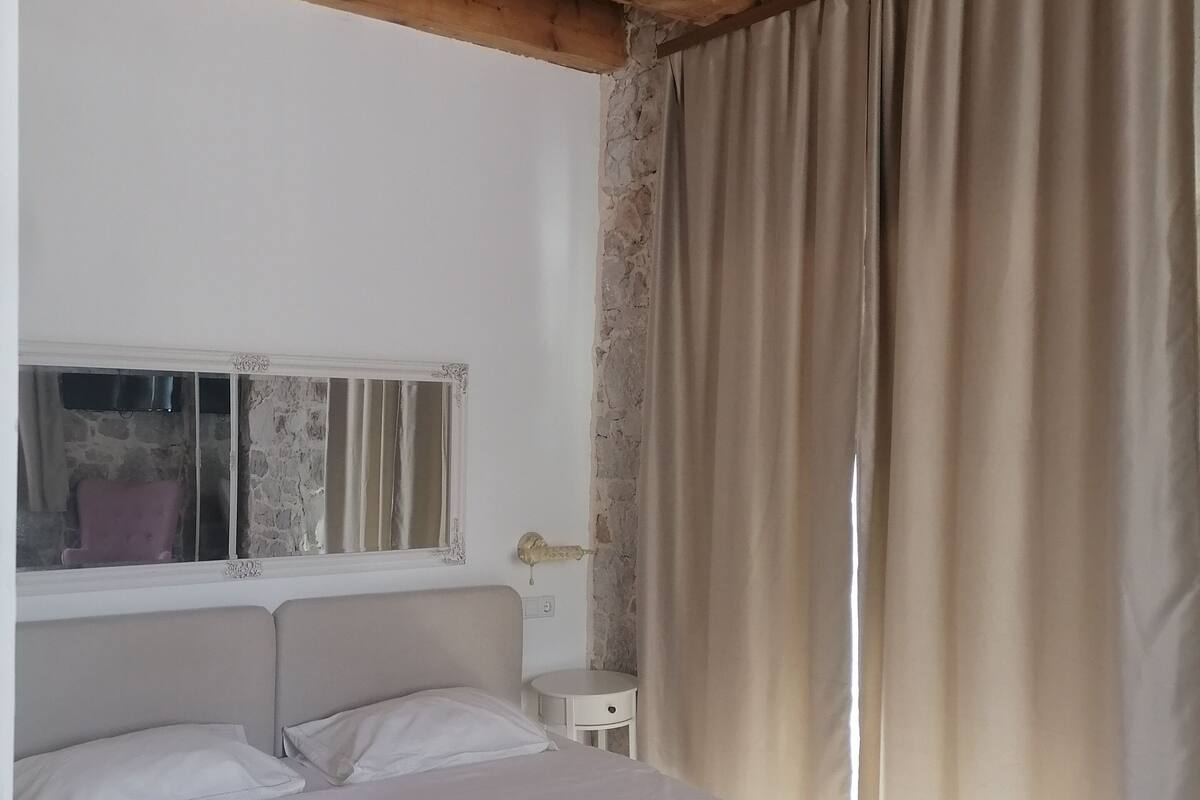 A bedroom with a wooden ceiling, a bed with a white bedspread, two rolled towels, a nightstand, a small round table, a window with a stone wall, and a curtain.