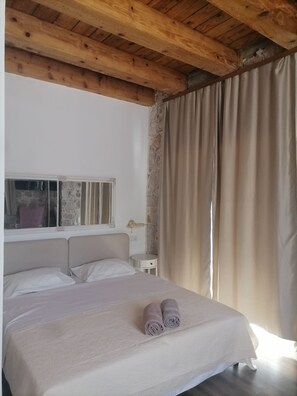 Une chambre avec un plafond en bois, un lit recouvert d’une courtepointe blanche, deux serviettes enroulées, une table de chevet, une petite table ronde, une fenêtre donnant sur un mur en pierre et un rideau.