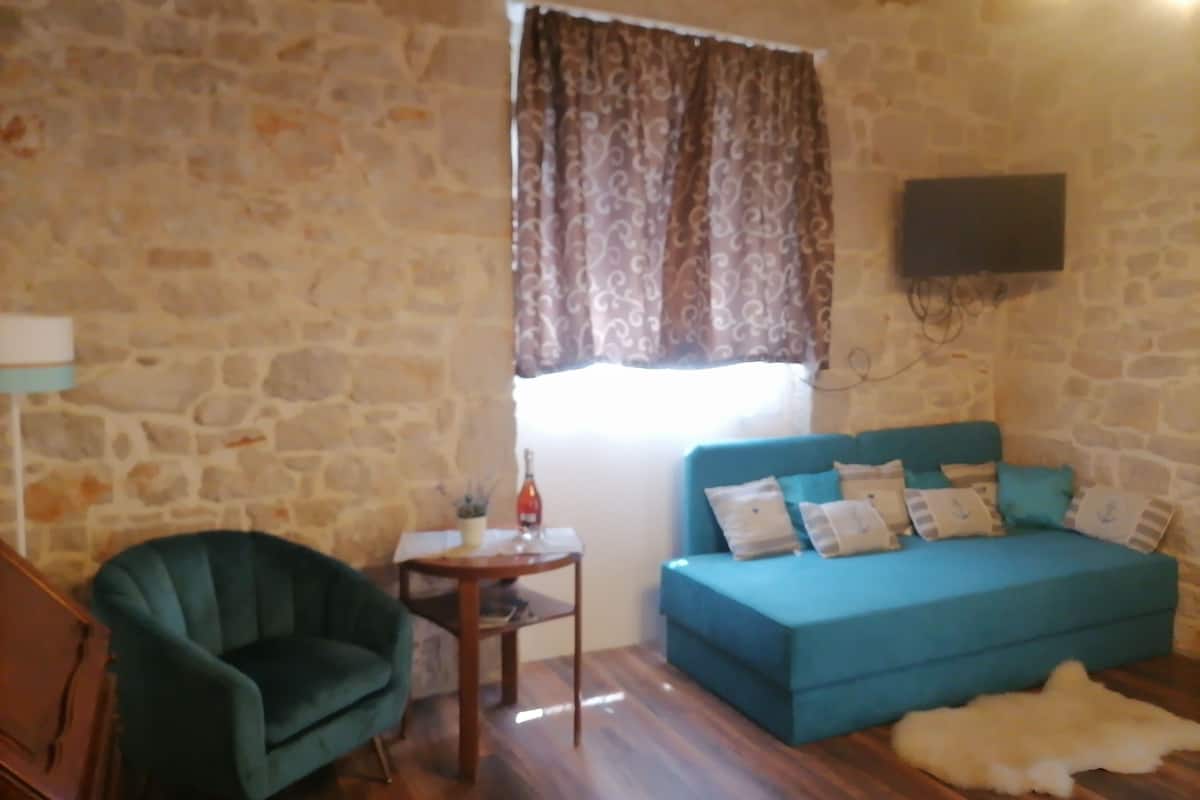 A room with a stone wall, a teal sofa with patterned cushions, a small wooden table with a bottle and a plant, a green armchair, and a white fur rug on the floor.
