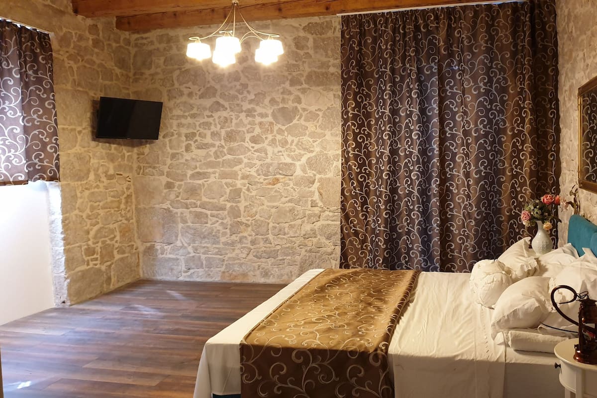 A bedroom with a stone wall, a wooden ceiling, a bed with a patterned bedspread, curtains with a swirl design, a mounted TV, and a bedside table with a lamp and a vase of flowers.