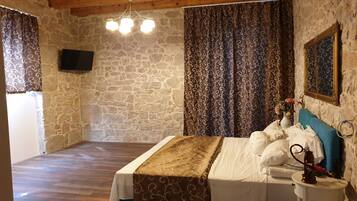 A bedroom with a stone wall, a wooden ceiling, a bed with a patterned bedspread, curtains with a swirl design, a mounted TV, and a bedside table with a lamp and a vase of flowers.
