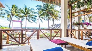 Un salón junto a la playa con barandillas de madera, sillones blancos y vistas a palmeras y al océano.
