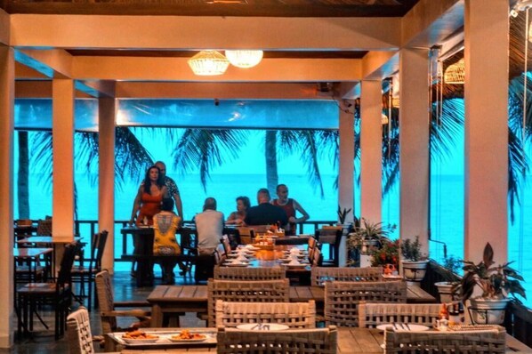 An indoor dining area with tables and chairs, a view of palm trees, and a ceiling with hanging lights.