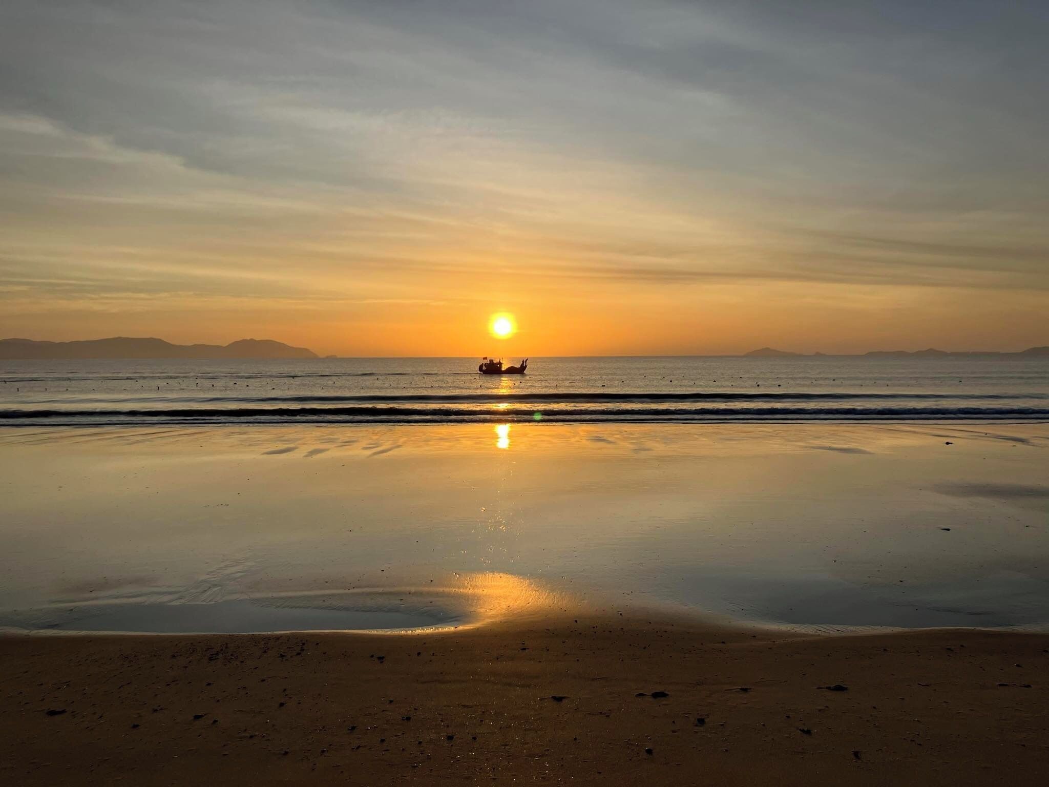 A beach at sunset with calm waters and a boat in the distance.