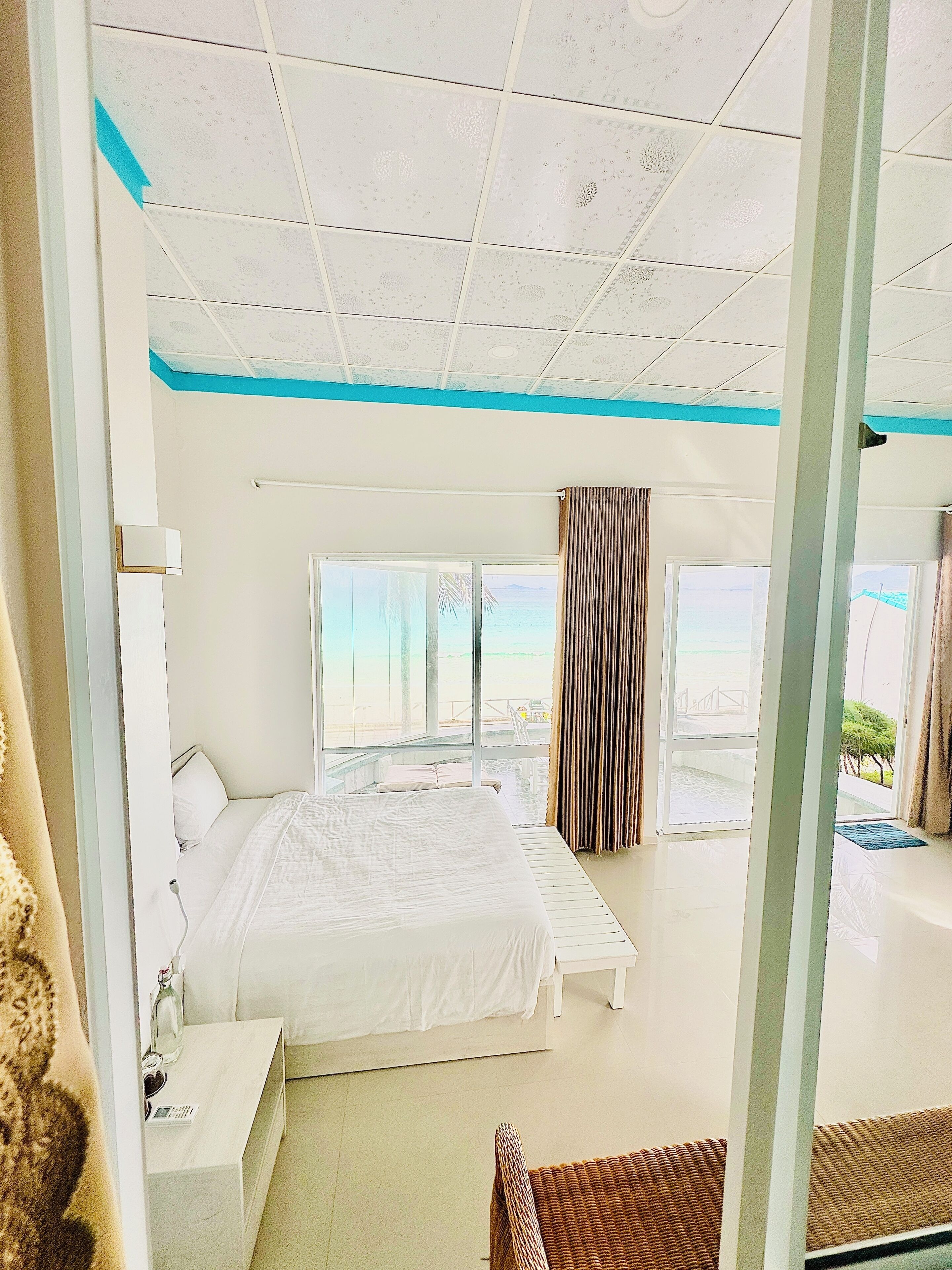 A hotel room with a bed, a bedside table, a window with curtains, and a view of the beach.