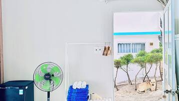 A room with a white air conditioner, a standing fan, a shelf with towels and slippers, and a view of a building and trees outside.