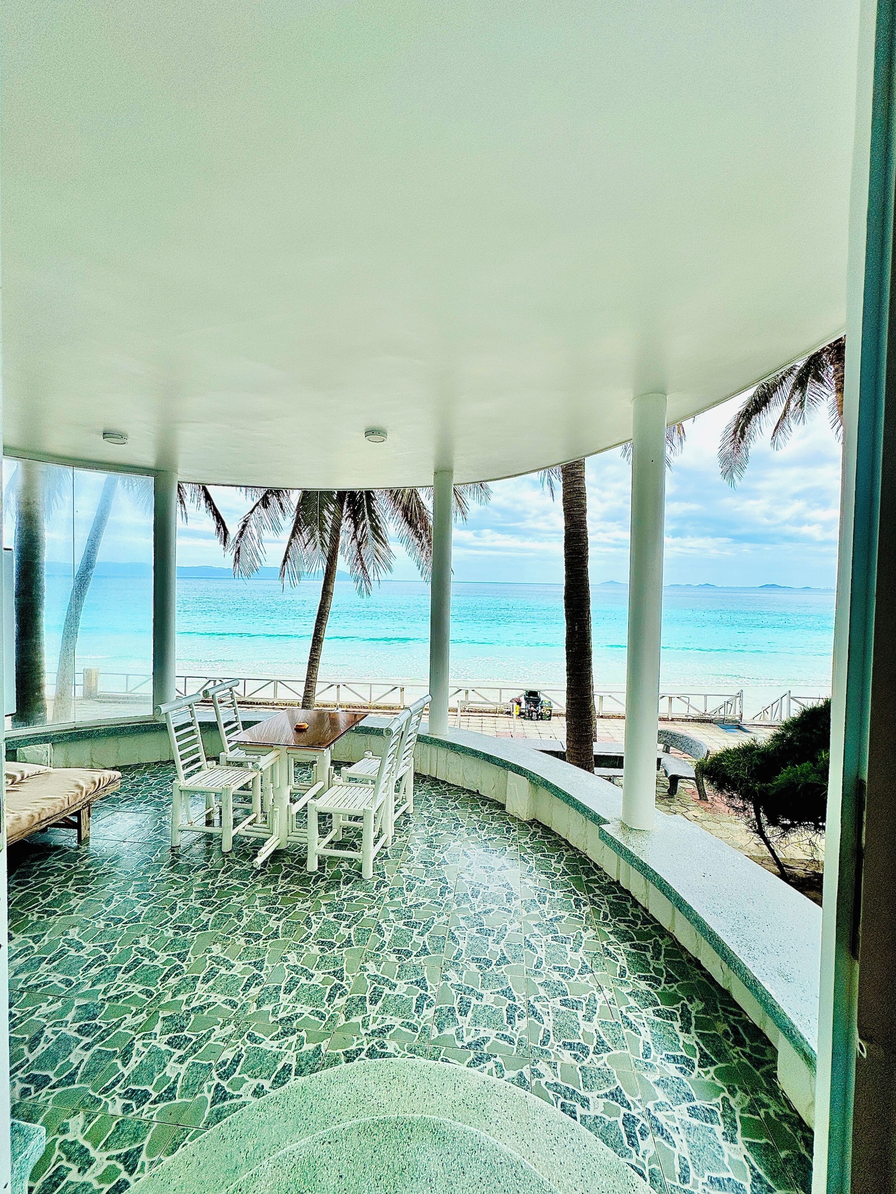 A covered outdoor seating area with a view of the ocean, featuring a mosaic floor and white furniture.
