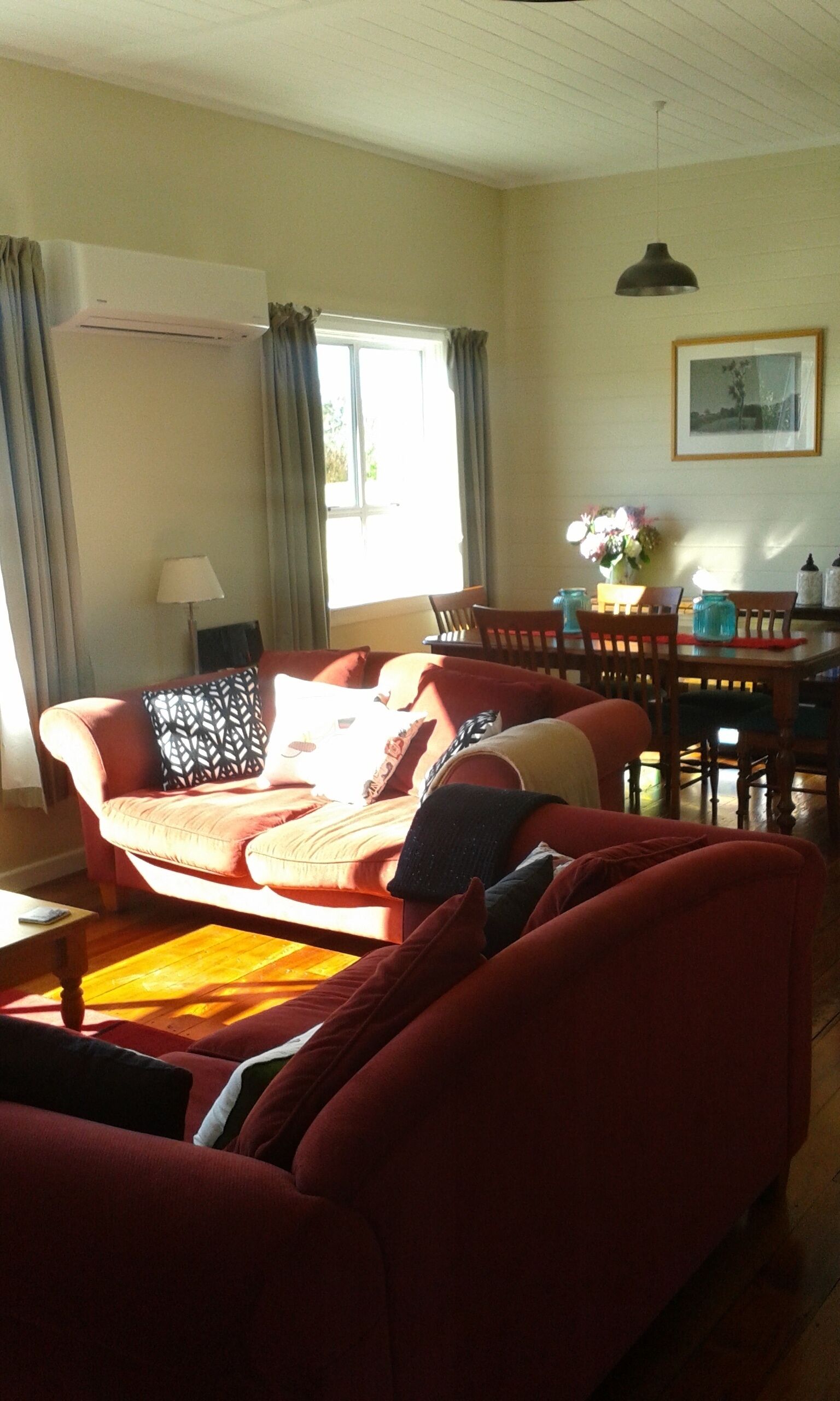 Ein Wohnzimmer mit einem roten Sofa, einem Esstisch mit Stühlen, einem Fenster mit Vorhängen und einem gerahmten Bild an der Wand.