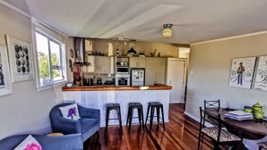 A modern kitchen with a central island, bar stools, a dining table with chairs, and a sofa with a decorative pillow. - Olive Tree Cottage, Martinborough (Martinborough)