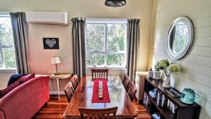 A dining area with a wooden table, chairs, and a red runner. There is a sofa, a lamp, and a bookshelf with books and decorative items. - Olive Tree Cottage, Martinborough (Martinborough)