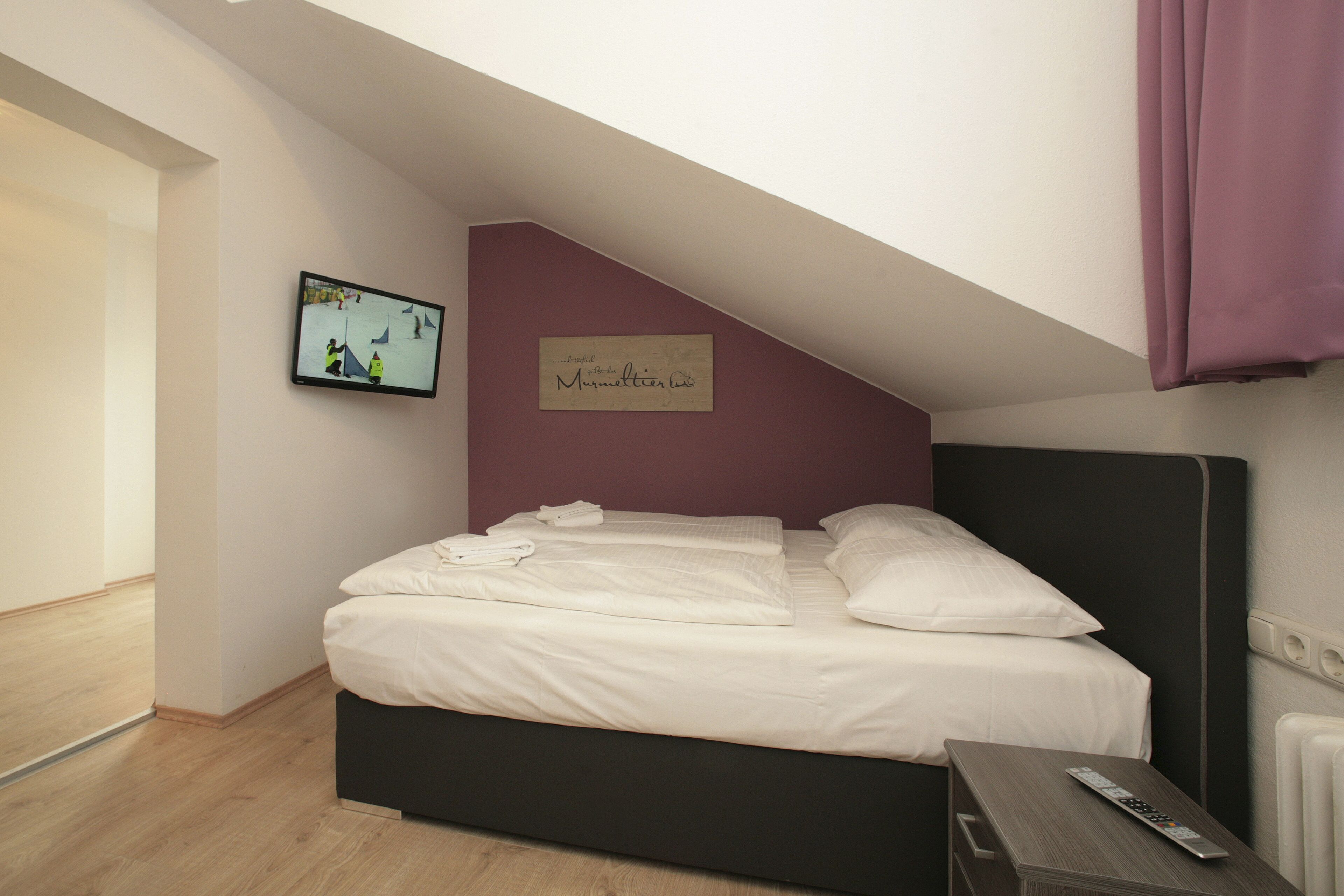 A bedroom with a bed, a bedside table, a remote control, and a wall-mounted TV.