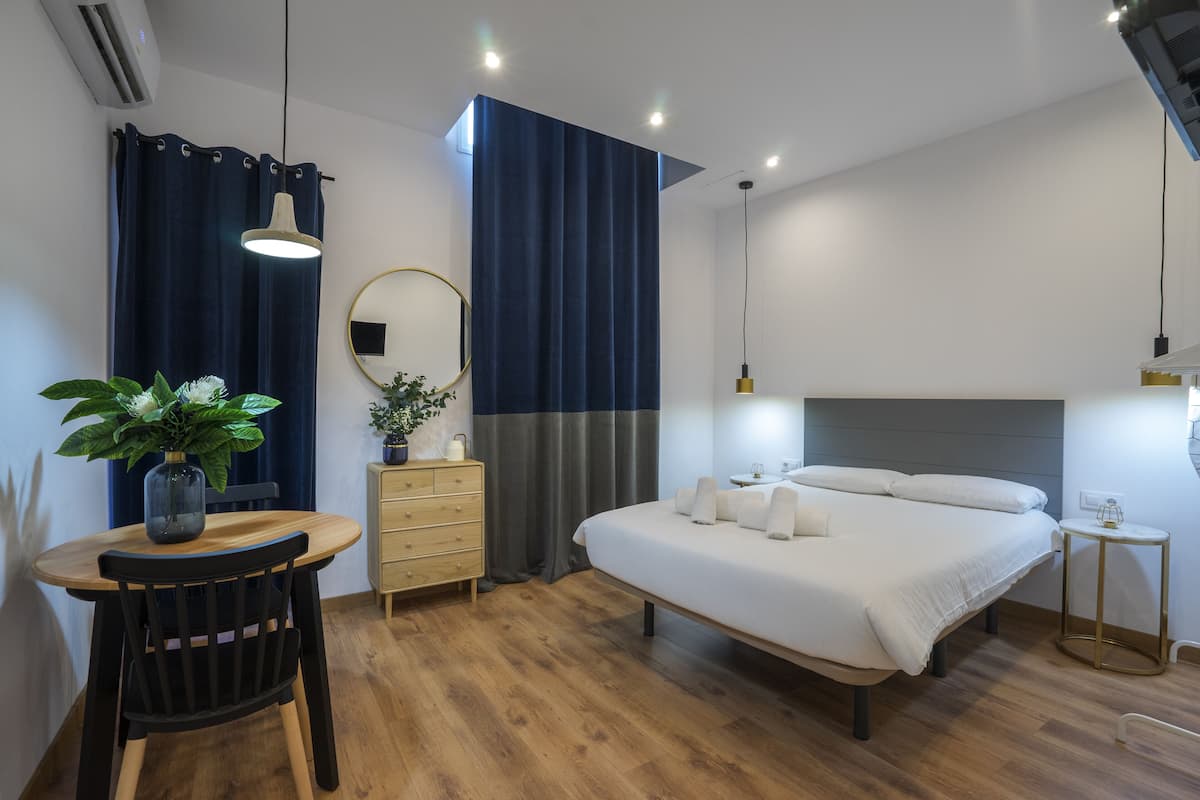 Quarto moderno com piso de madeira, uma cama com lençóis brancos, mesa de cabeceira, cômoda, espelho e um vaso com planta.