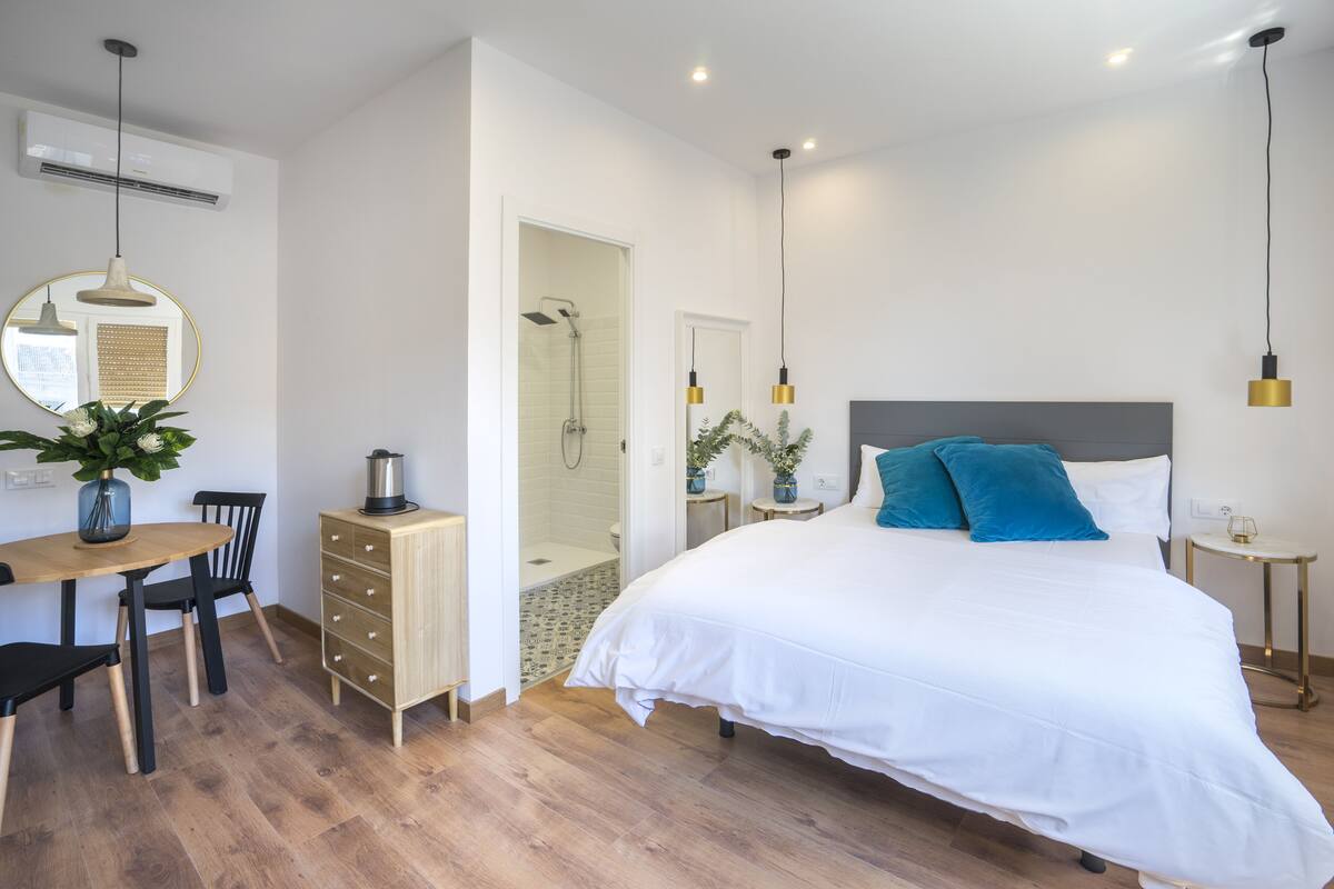 Quarto moderno com cama grande, mesas de cabeceira, escrivaninha com cadeira, área de chuveiro e pequena mesa com uma planta.