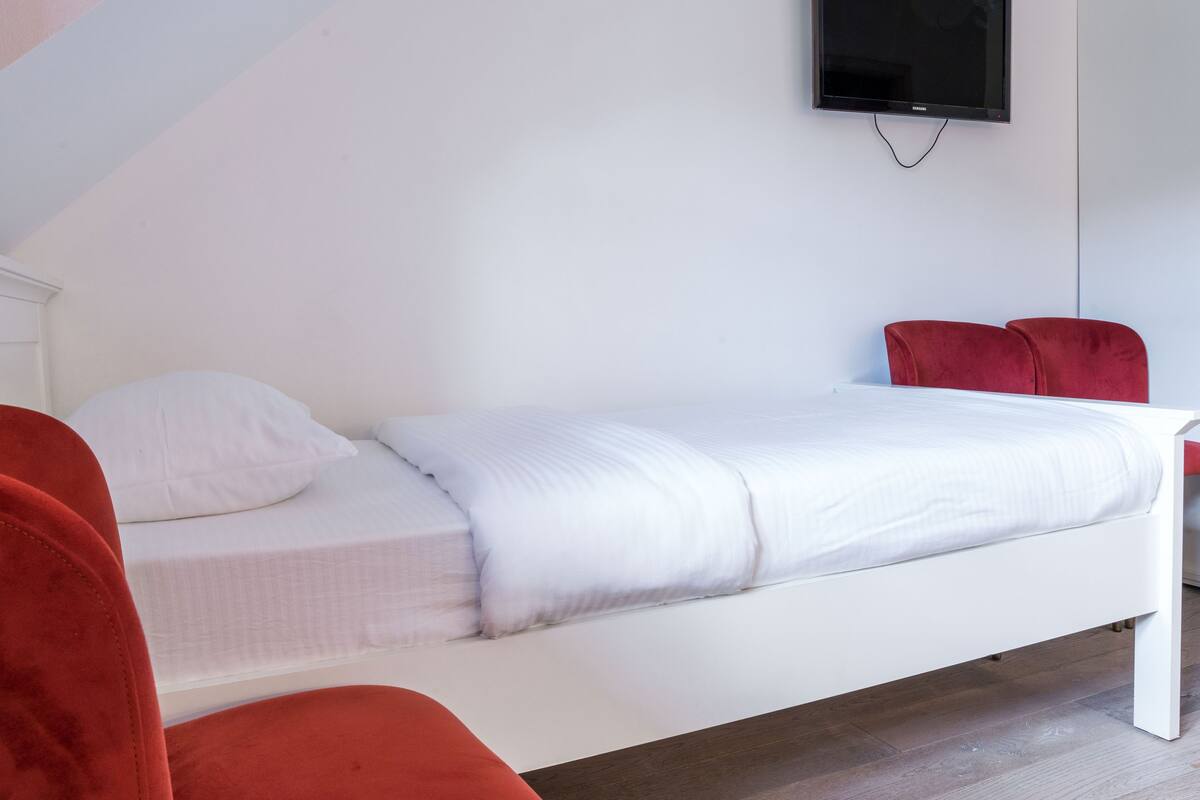 Una cama individual con ropa de cama blanca, una silla roja y un televisor montado en la pared.