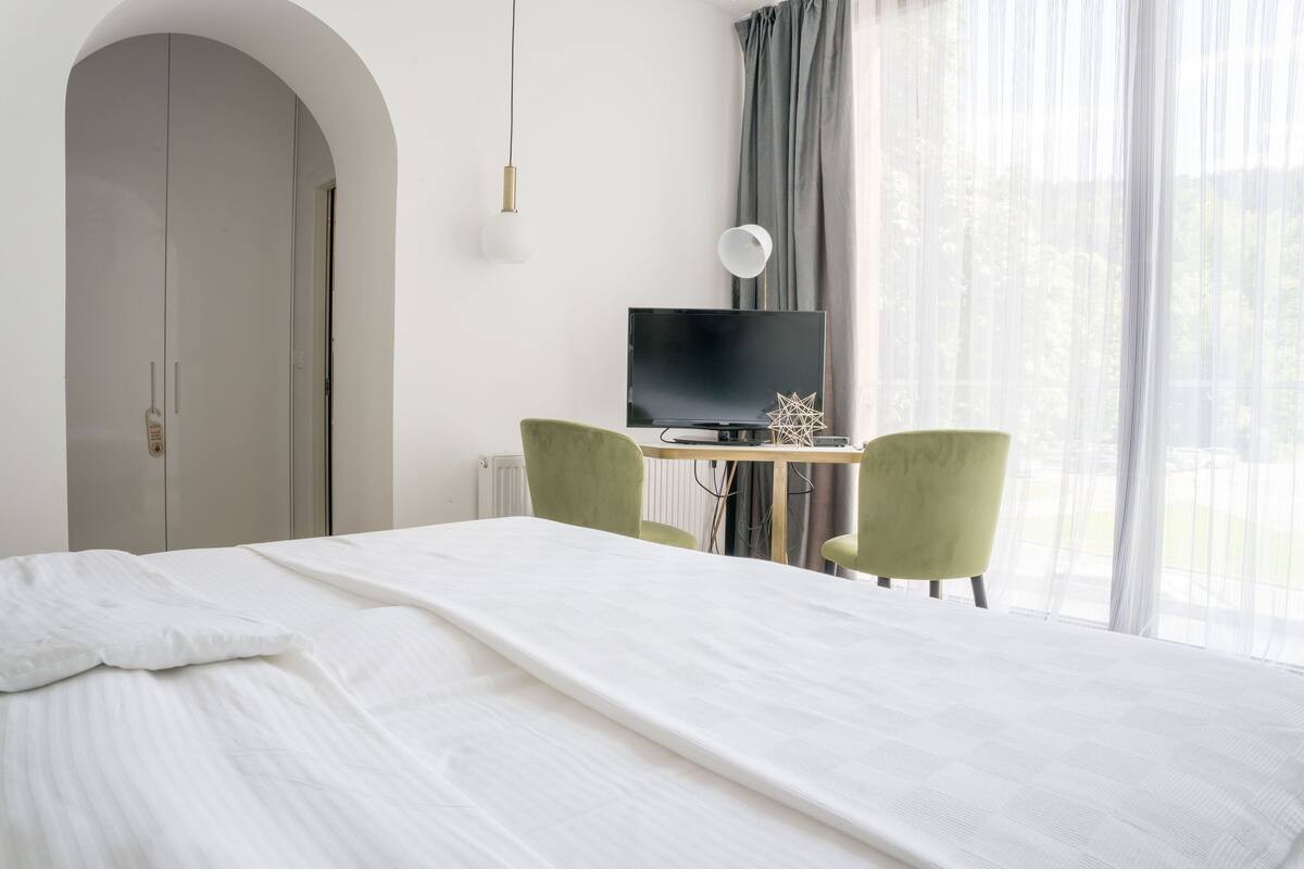 Habitación de hotel con cama, escritorio con computadora, dos sillas, televisión y ventana con cortinas.