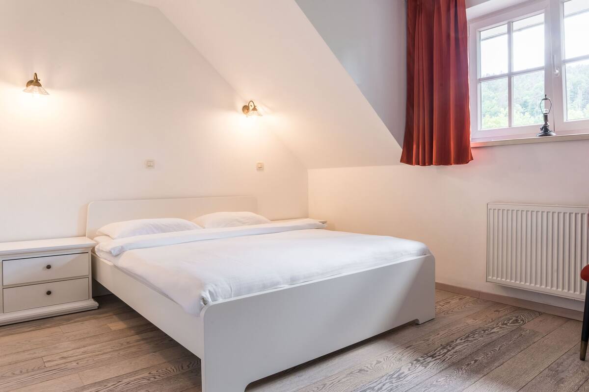 Un dormitorio con una cama blanca, dos mesitas de noche, un ventanal con cortina roja y un radiador.