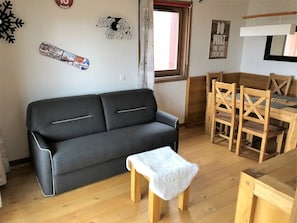 A compact living space with a black sofa, a small wooden stool, a dining table with wooden chairs, and a window with curtains.
