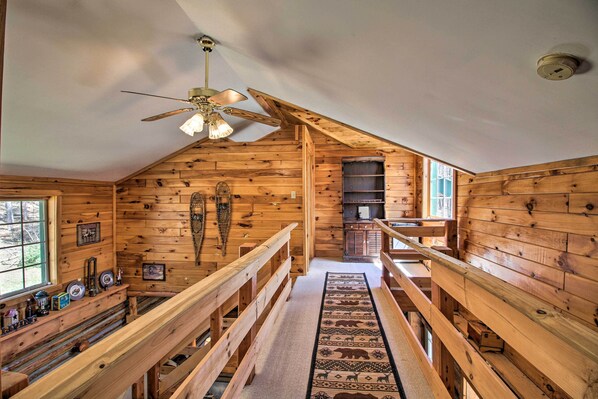 Interior de una cabaña de madera con techo alto, una escalera central y un ventilador de techo.