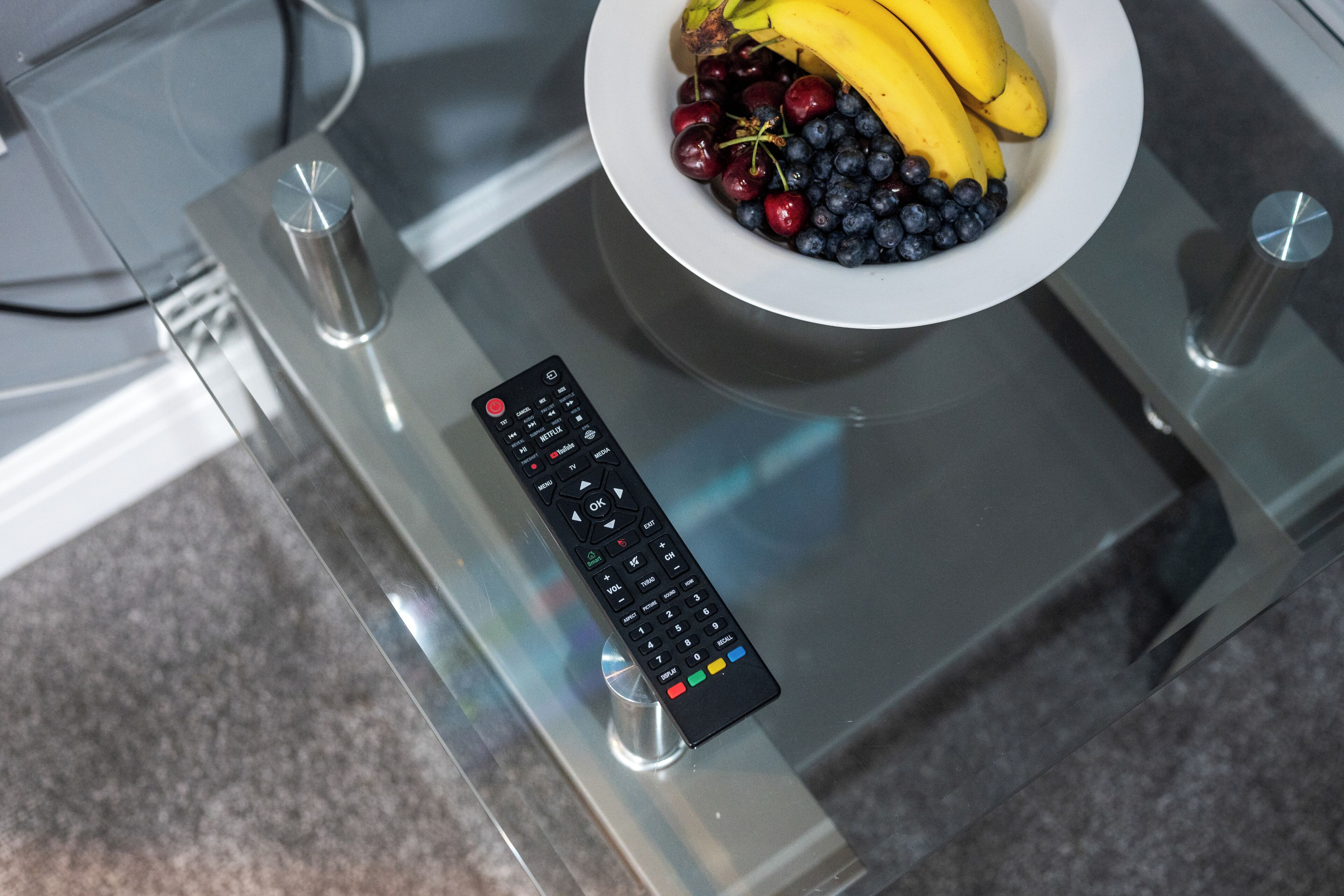 A remote control on a glass table with a bowl of fruit.