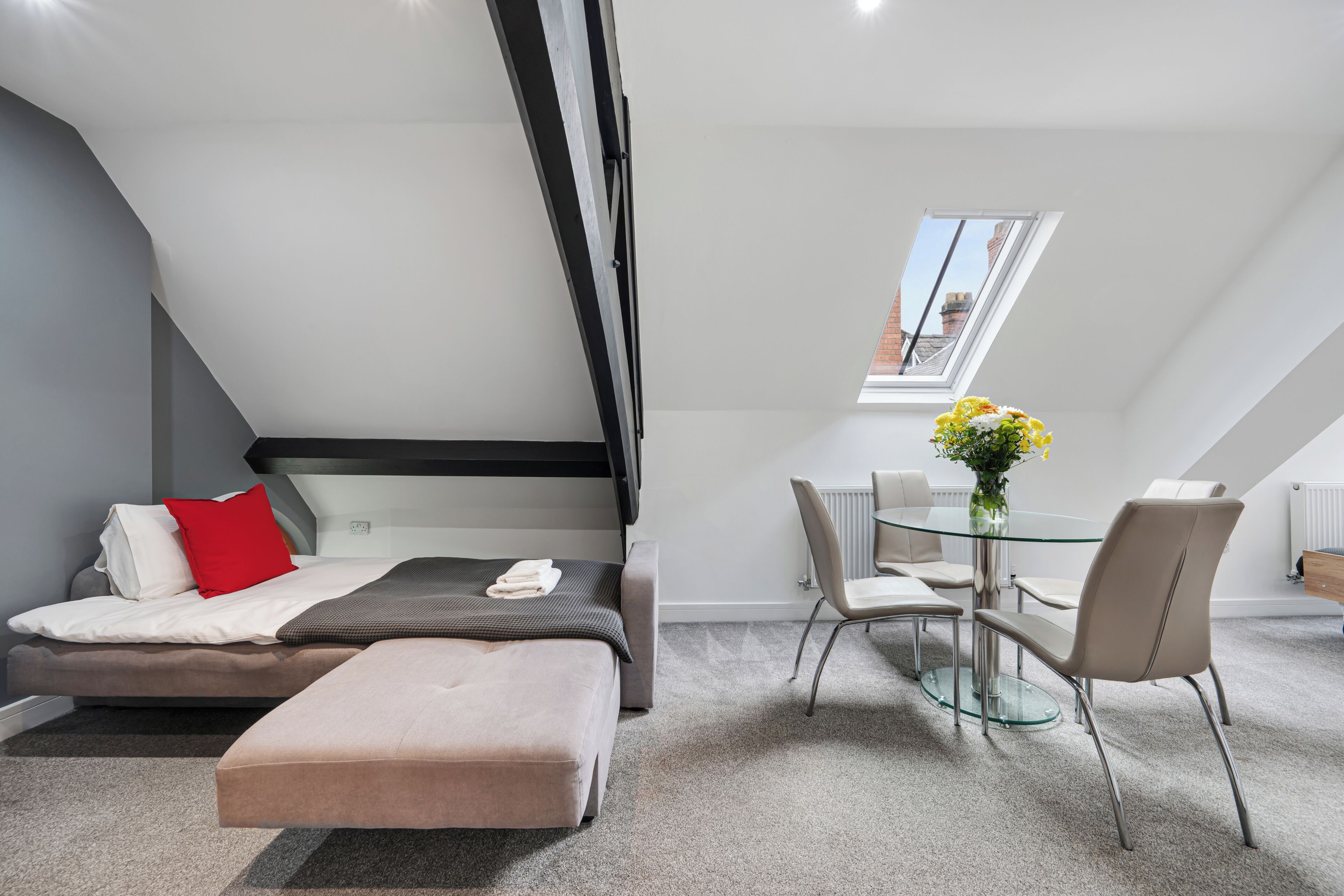 A modern room with a bed, a dining table with chairs, a small window, and a skylight.
