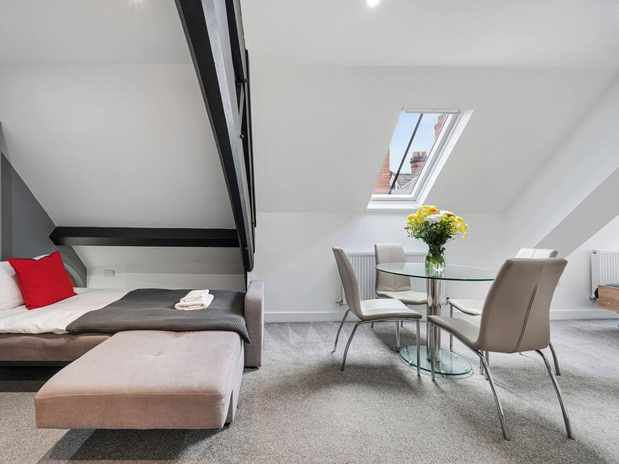 A modern room with a bed, a dining table with chairs, a small window, and a skylight.