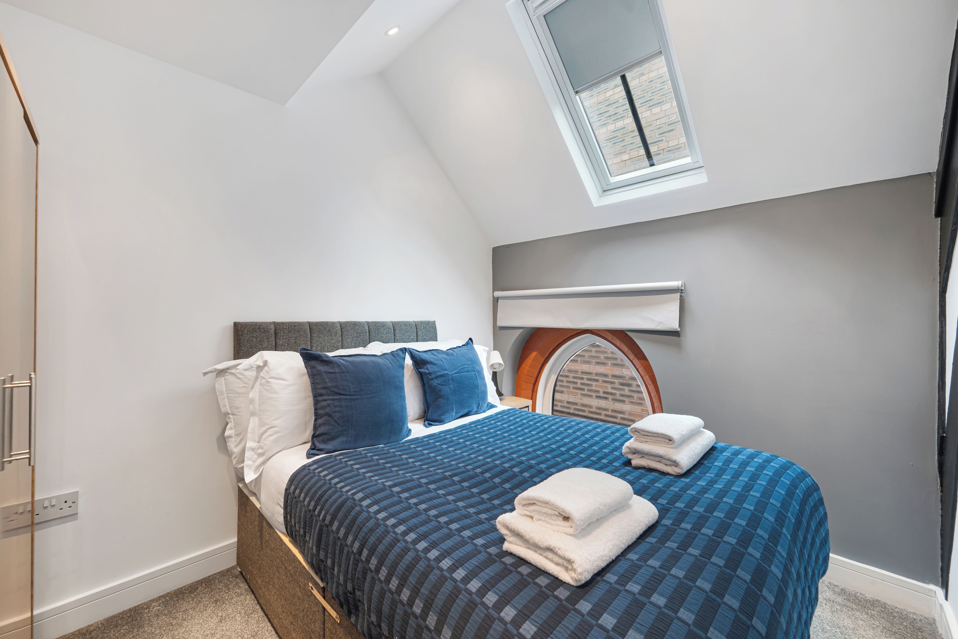 Un dormitorio con cama, cabecera, ventana y una lámpara fijada a la pared.