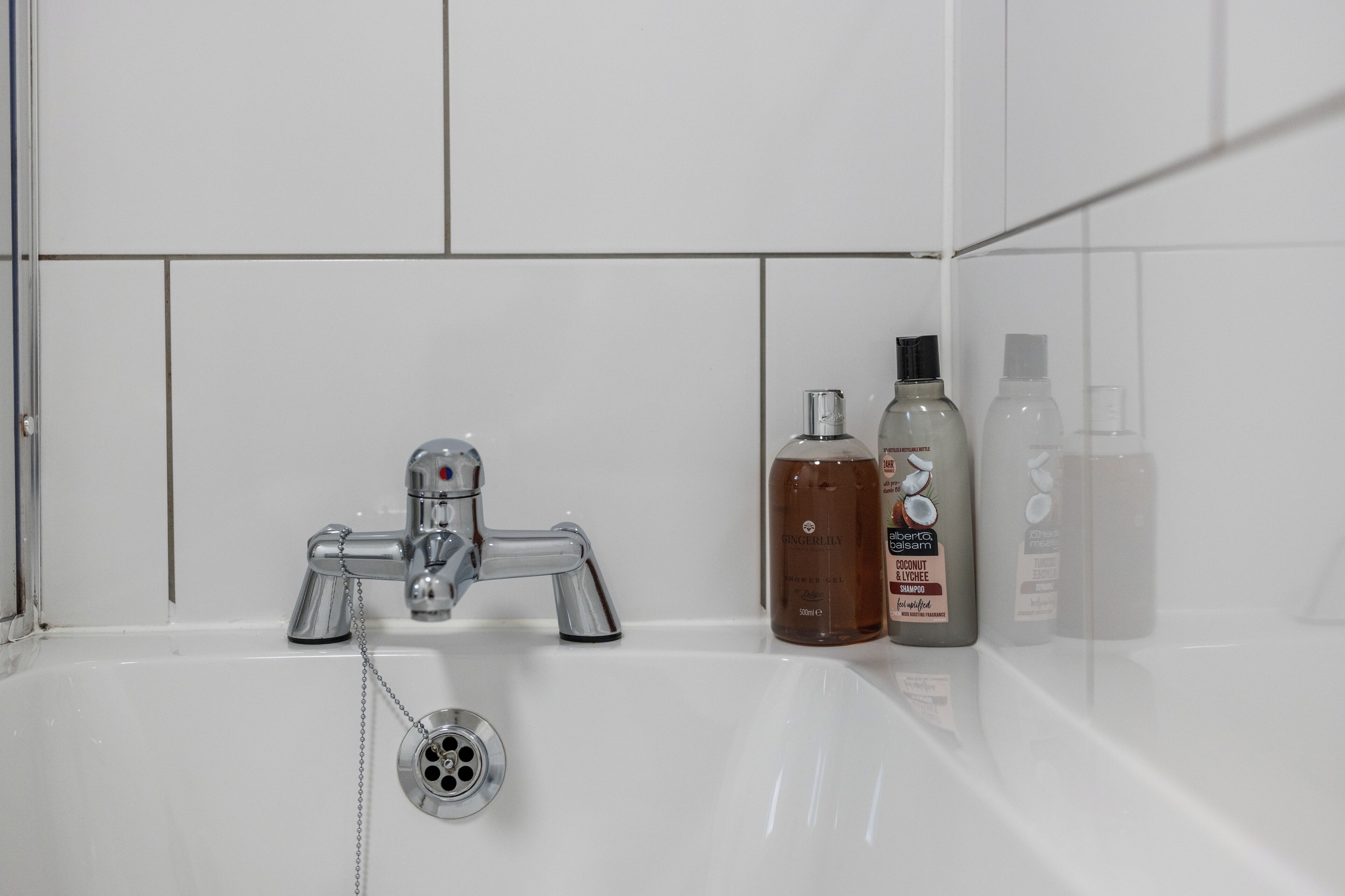 Un baño con pared de azulejos blancos, grifo cromado y tres botellas de artículos de tocador sobre el lavamanos.