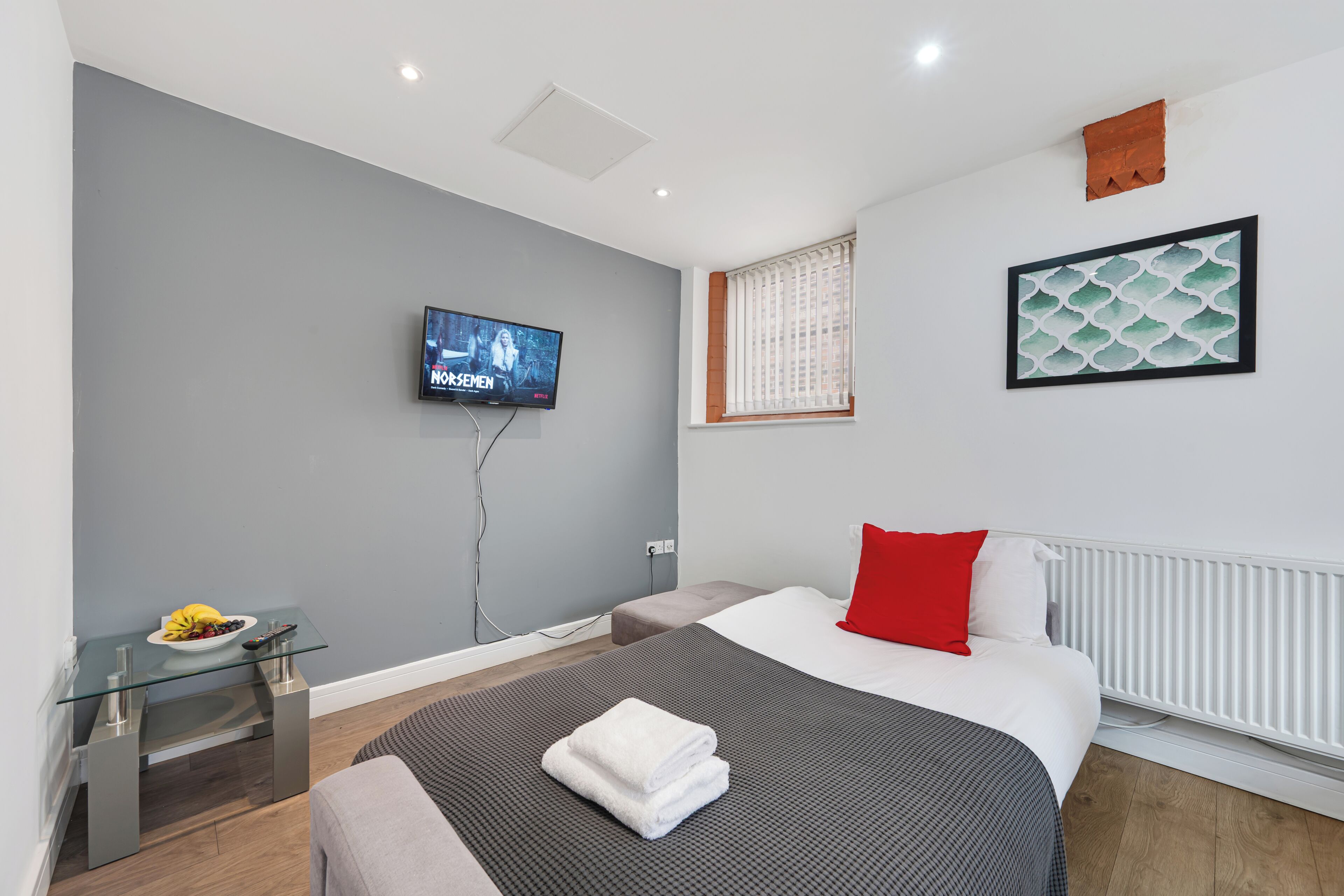 A modern hotel room with a bed, a TV, a small table with fruit, and a window with blinds.