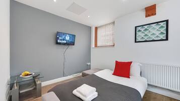 A modern hotel room with a bed, a TV, a small table with fruit, and a window with blinds.