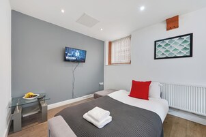 A modern hotel room with a bed, a TV, a small table with fruit, and a window with blinds.