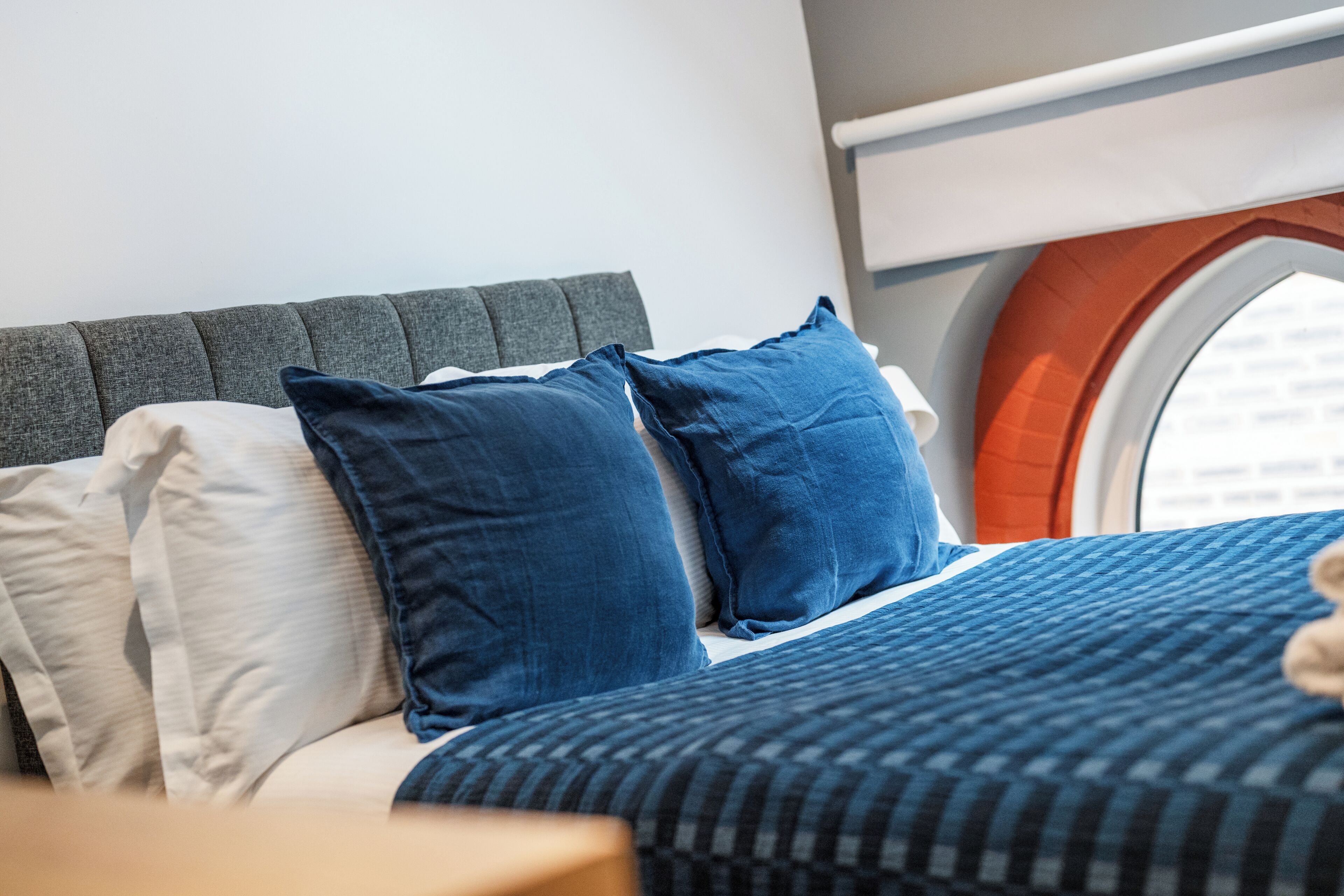 Uma cama com cabeceira estofada cinza, almofadas azuis e brancas, e um edredom listrado em azul e branco.