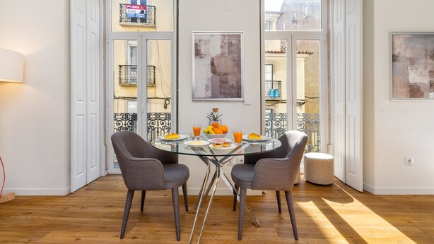 Un comedor con una mesa de vidrio, cuatro sillas grises y una pequeña mesa redonda con fruta y bebidas.