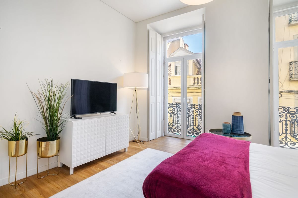 Ein modernes Schlafzimmer mit einem Bett, einem Fernseher, einer weißen Kommode und zwei Zimmerpflanzen.