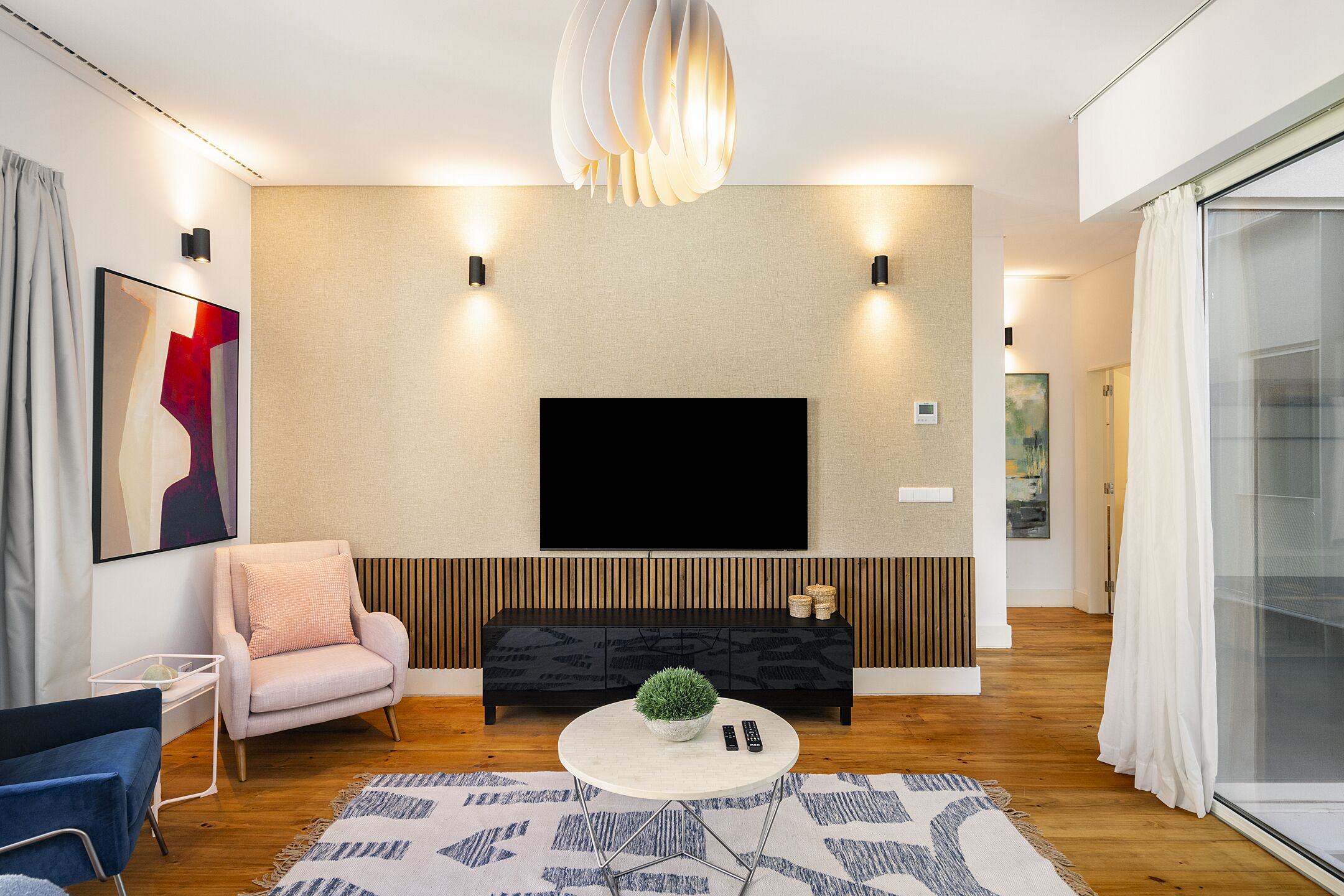 A modern living room with a flat-screen TV mounted on a textured wall, a round coffee table with a plant, a pink armchair, and a blue sofa.