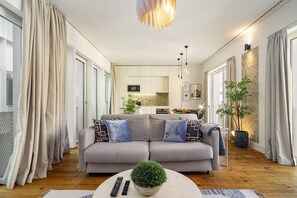Uma sala de estar moderna com um sofá cinzento, uma mesa de centro com uma planta e uma cozinha ao fundo.