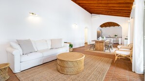 A modern living room with a white sofa, a round wicker ottoman, and a dining area with a wooden table and chairs. - Comporta Family Beach House (Alcacer do Sal)