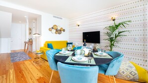 Un coin repas avec une table ronde dressée pour quatre personnes, entourée de chaises bleues. Un canapé jaune et une petite table d’appoint avec une lampe sont visibles à l’arrière-plan.