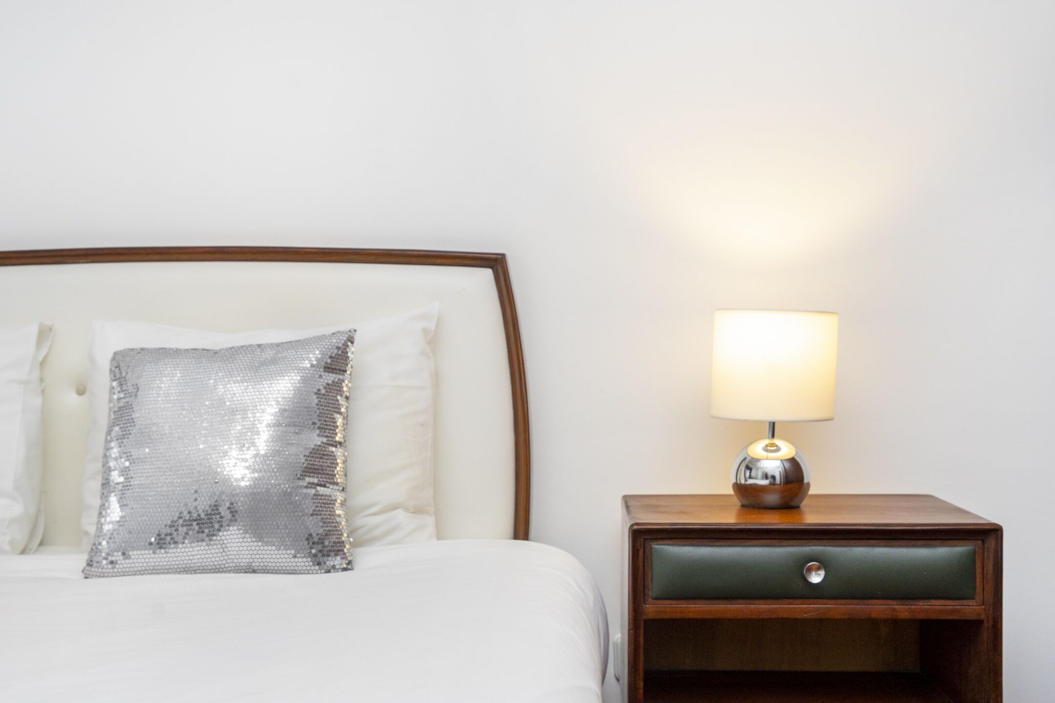 A neatly made bed with a sequined pillow, a bedside lamp, and a wooden nightstand with a drawer.