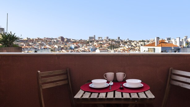 Ein Tisch mit einer roten Tischdecke, zwei weißen Schüsseln und zwei weißen Tassen auf einem Balkon mit Blick auf eine Stadtlandschaft.