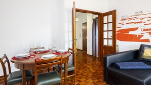 Ein Essbereich mit einem runden Holztisch, gedeckt für vier Personen, umgeben von Holzstühlen mit grünen Kissen. Gegen eine Wand mit einem großen orangefarbenen und weißen Wandgemälde ist eine schwarze Ledersofa mit blauer Decke gestellt.