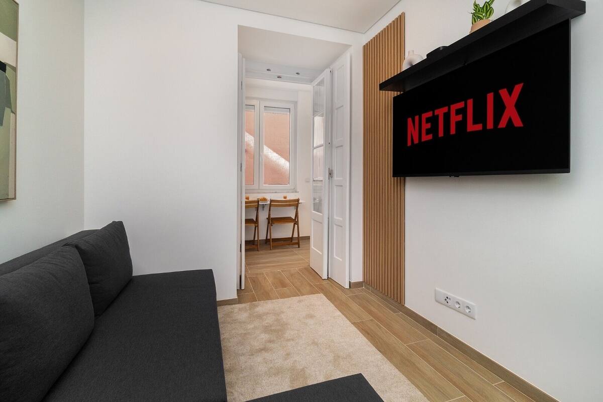 A modern living room with a grey sofa, a wooden floor, a carpet, and a wall-mounted TV displaying the Netflix logo.