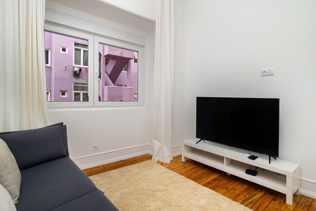 A living room with a sofa, a TV on a white stand, a window with a view of a building, and a wooden floor.