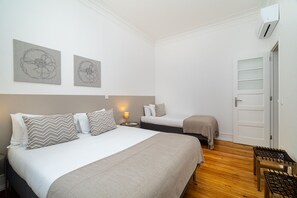 Una habitación de hotel con una cama grande, dos camas más pequeñas, una mesita de noche con lámpara, un banco de madera y una obra de arte en la pared.