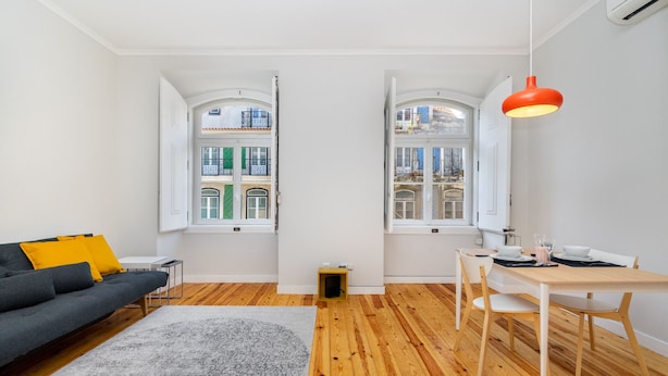 Una sala de estar moderna con un sofá, una mesa de comedor con sillas, un tapete y una ventana con contraventanas abiertas.