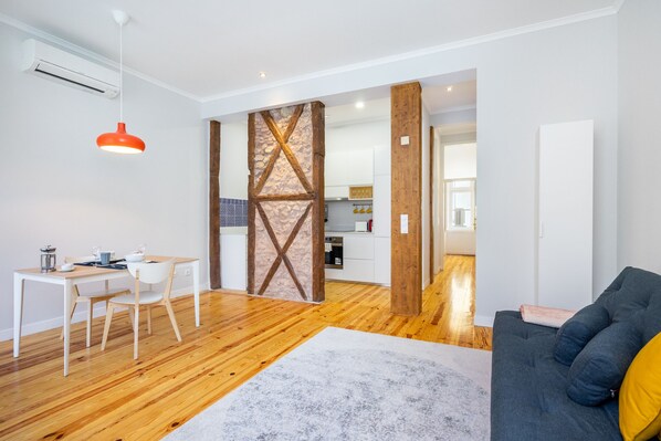 Una sala de estar moderna con suelo de madera, un sofá gris, una mesa de comedor con sillas y una cocina al fondo.