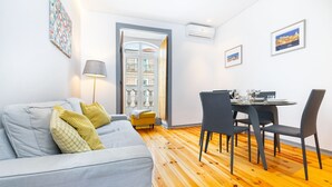 Ein Wohnzimmer mit einer Couch, einem Esstisch mit Stühlen, einer Lampe und Blick durch eine Tür.