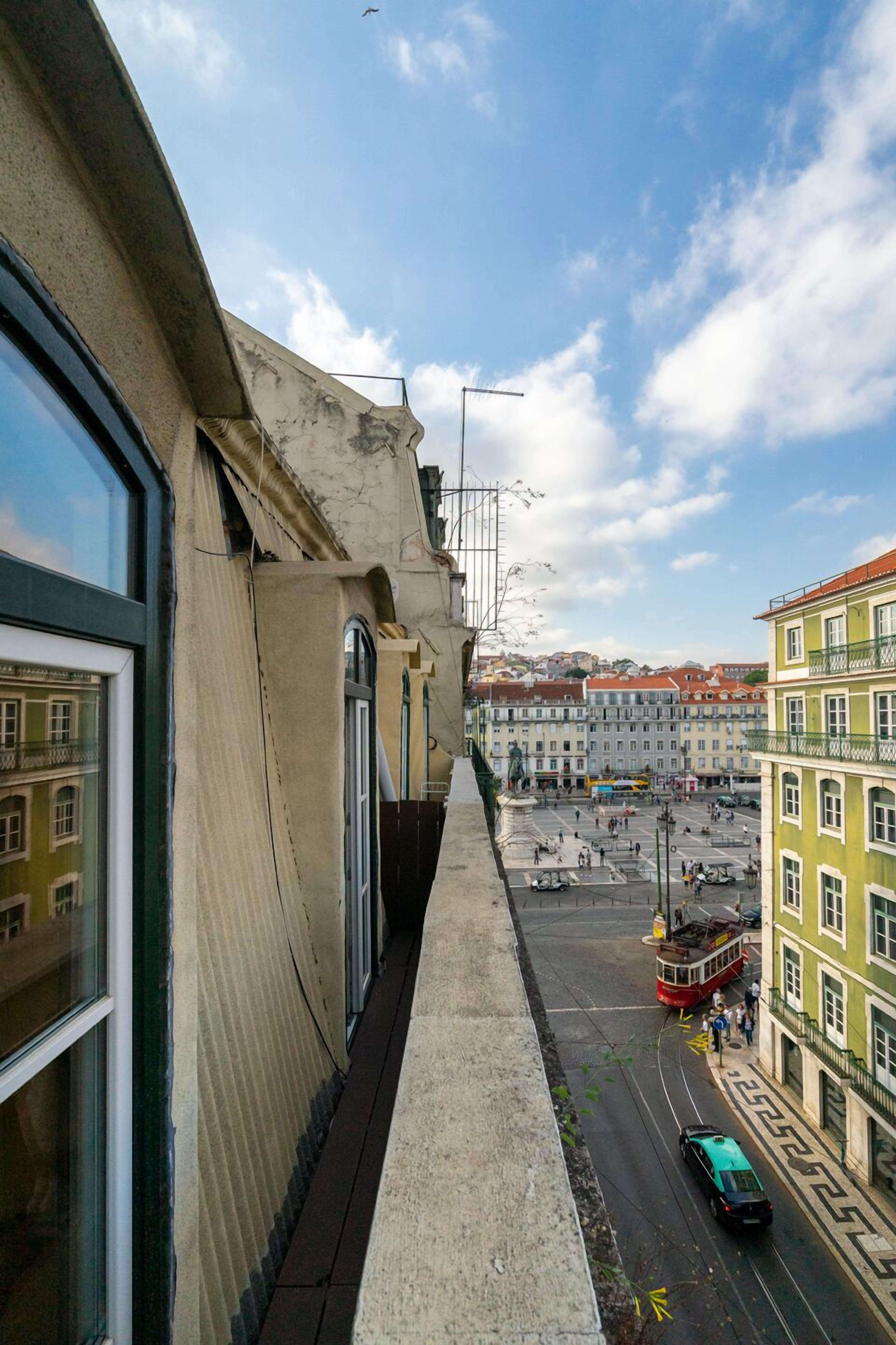A narrow urban street with a tram and a car, buildings with balconies, and a clear sky.