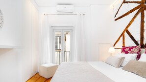 Ein Schlafzimmer mit Holzboden, einem Bett mit weißer Bettdecke, einem Nachttisch mit Lampe und Blick auf einen Balkon durch eine Tür.