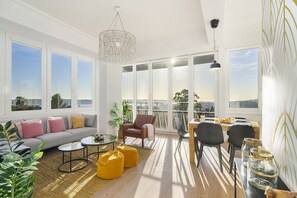 Amplia sala de estar con sofá, mesa de centro y sillones, ventanas grandes con vistas y un balcón con barandilla.