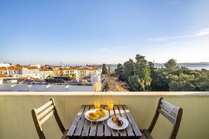 Un balcon avec une table en bois mise pour le petit-déjeuner, d’où l’on peut admirer un paysage urbain et une étendue d’eau.